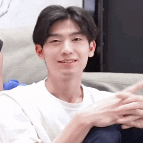 a young man in a white shirt is sitting on a couch with his hands folded and smiling .