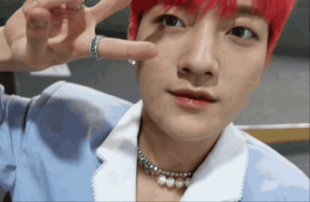 a young man with red hair and a pearl necklace is making a peace sign with his hand .