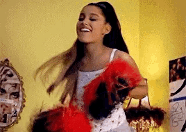 ariana grande is wearing a white tank top and holding a red and black feather boa while dancing in a room .