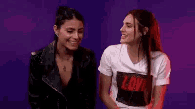 two women are sitting next to each other in front of a purple background and smiling .