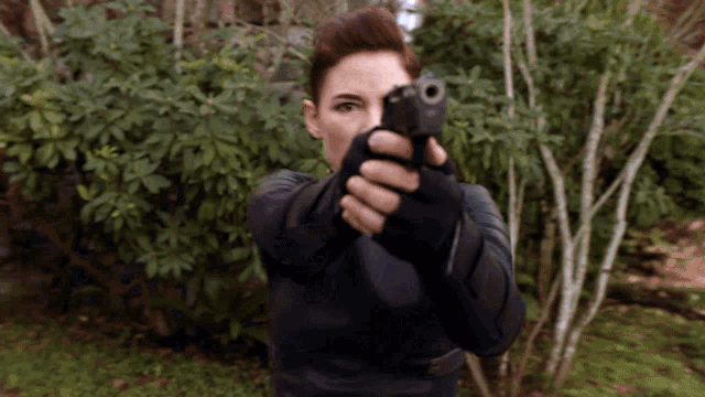 a woman is pointing a gun at the camera with trees in the background