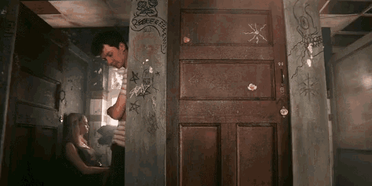 a man standing next to a woman in a room with graffiti on the walls including one that says queer free