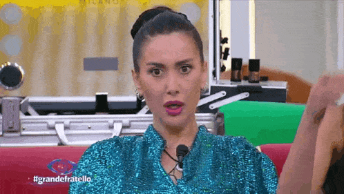 a woman wearing a blue sequined top is sitting on a red couch talking into a microphone .