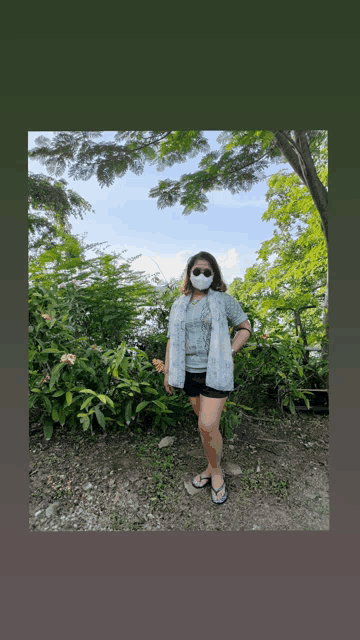 a woman wearing a mask and flip flops stands in front of a bush