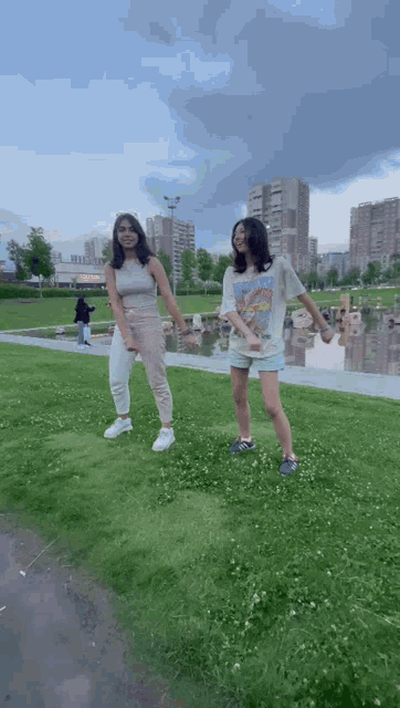 two girls are dancing in a park in front of a pond .