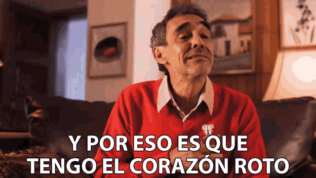 a man in a red sweater sits on a couch with the words y por eso es que tengo el corazon roto below him
