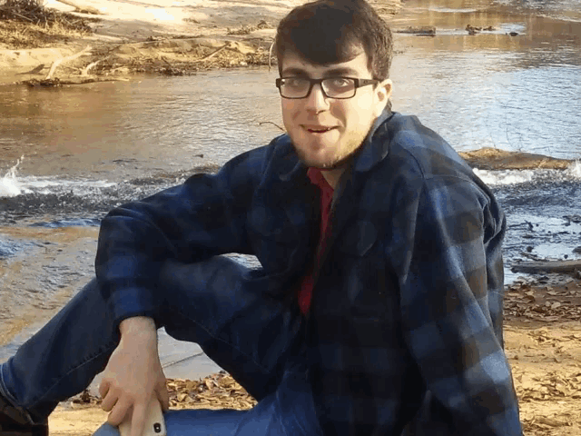 a man wearing glasses and a plaid shirt sits on the ground near a river