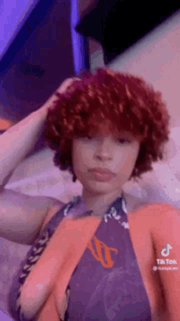 a woman with red curly hair is sitting on a bed with her hand on her hair .