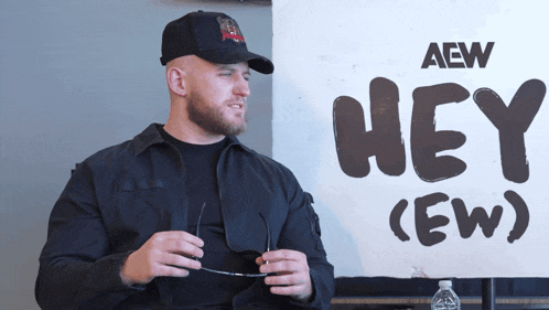 a man in a black jacket stands in front of a sign that says hey ( ew )