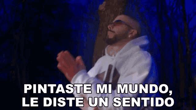 a man with a beard and sunglasses is standing in front of a tree with his hands folded in prayer .