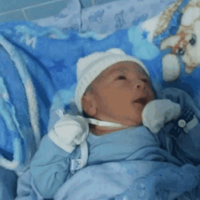 a baby wearing a hat and gloves is laying on a blue blanket .