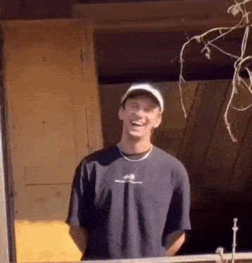 a man wearing a hat and a black t-shirt is laughing while standing in front of a wooden door .