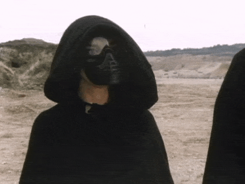a person wearing a black hooded cape and a black mask is standing in a dirt field .