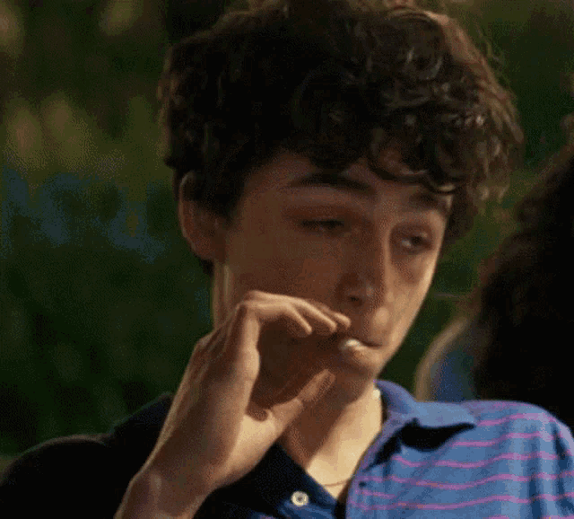 a young man with curly hair is smoking a cigarette while wearing a blue striped shirt .