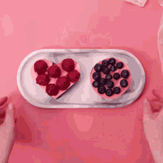 a heart shaped cake and a bowl of blueberries on a tray on a pink table .
