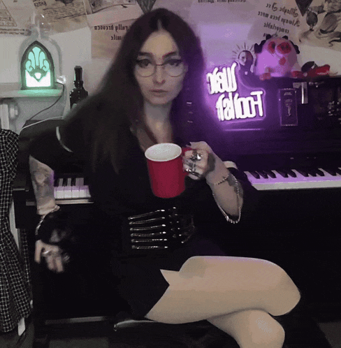 a woman sitting in front of a piano with a red cup in her hand and a neon sign that says your fatboot