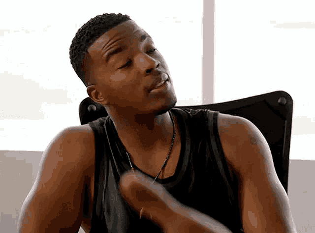 a man in a black tank top is sitting in a chair and making a funny face .