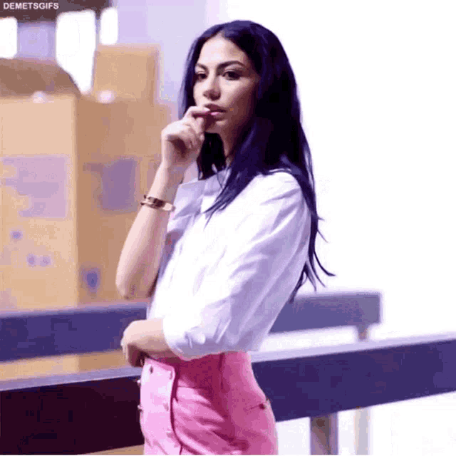 a woman in a white shirt and pink pants is standing next to a railing with her hand on her chin .