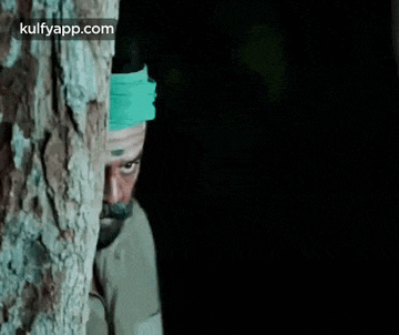 a man with a mustache and a green headband is standing next to a tree .
