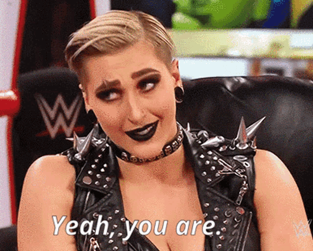 a woman in a leather vest with spikes on it is sitting in a chair and saying yeah you are .