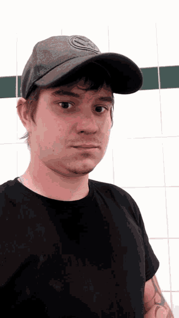 a man wearing a hat and a black shirt stands in front of a white wall