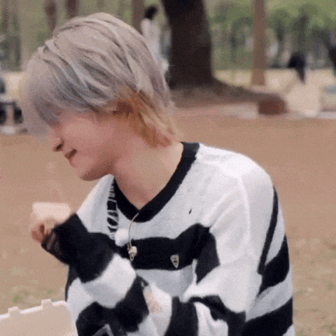 a young man wearing a black and white striped sweater is sitting on the ground eating a sandwich .