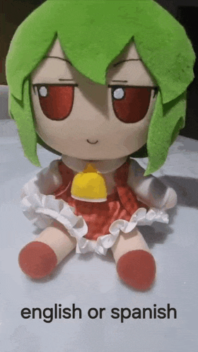 a stuffed doll with green hair and red eyes is sitting on a table next to the words english or spanish .
