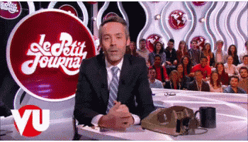 a man in a suit and tie sits at a table in front of a sign that says le petit journal