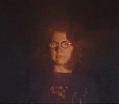 a woman wearing glasses and a black shirt is standing in the dark with her hands up .