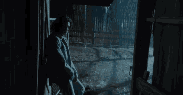 a man sits in a dark room looking out a window at the rain