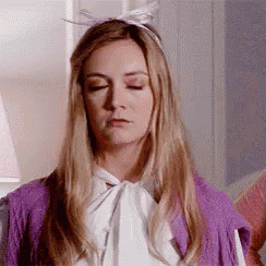 a woman wearing a purple cardigan and a white shirt with a bow tie is making a funny face .