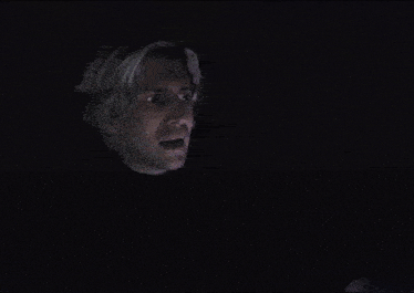 a man with blonde hair is looking at something in the dark and making a surprised face .
