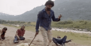 a man is walking across a dirt field with a stick while a group of people are sitting on the ground .