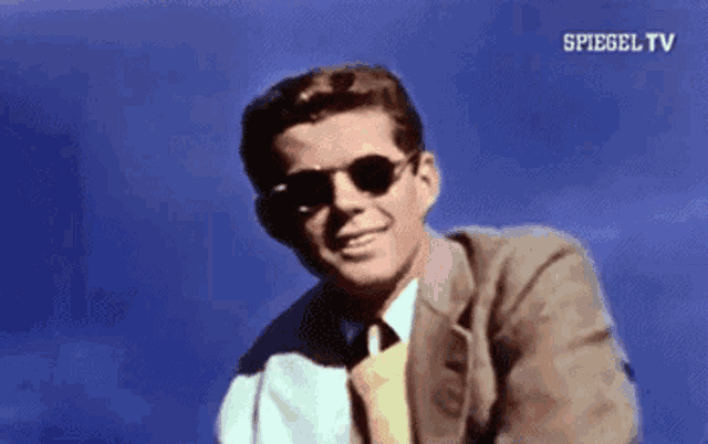 a man in a suit and tie is wearing sunglasses and smiling in front of a blue sky .