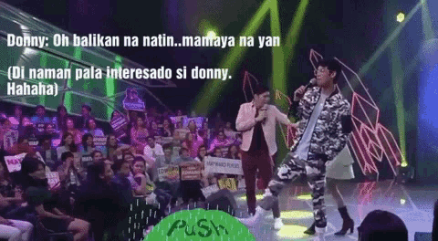a man in a camouflage jacket stands on a stage with a microphone in front of a crowd of people