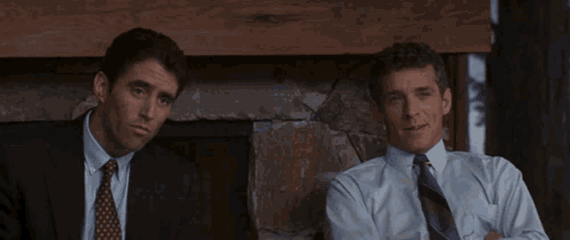 two men in suits and ties are sitting in front of a stone fireplace