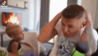 a man is sitting on a couch with his hands on his head and a child is playing with him .