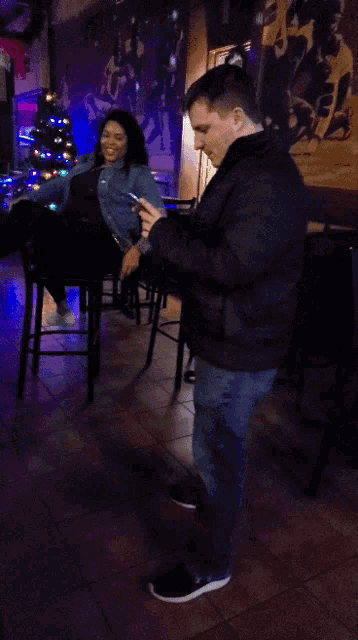 a man standing next to a woman sitting on a bar stool looking at her phone