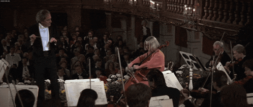 a woman in a pink dress is playing a cello in front of a large orchestra