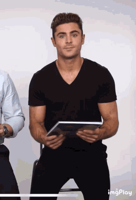 a man in a black t-shirt is holding a piece of paper with the word imgplay below him