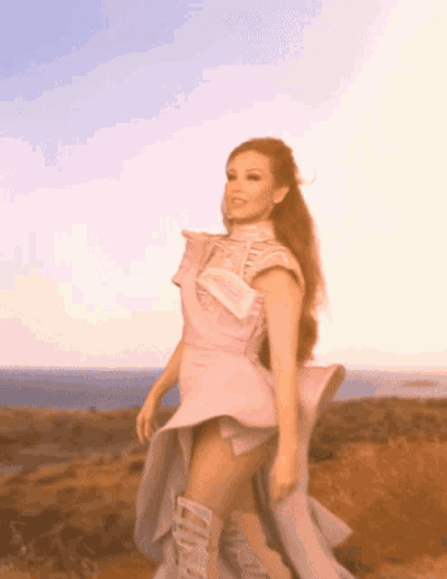 a woman in a pink dress and boots stands on a hill overlooking the ocean
