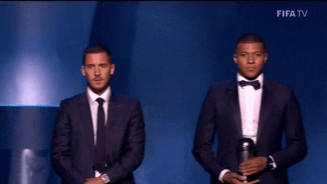 three men in suits and bow ties are on a stage with fifa tv in the upper right corner