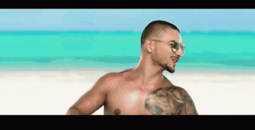 a shirtless man wearing sunglasses and a tattoo on his chest is standing on a beach .