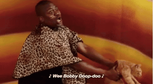 a man in a leopard print shirt is holding a cat and singing wee bobby doop-doo .