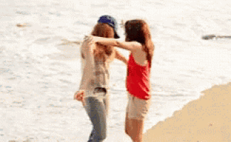 two women hugging on the beach with one wearing a hat that says ny