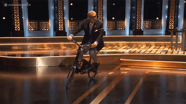 a man in a suit is riding a bicycle on a stage with the hashtag #cbcdragonsden