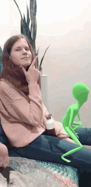 a girl in a pink sweater sits on a couch with a green alien on her lap