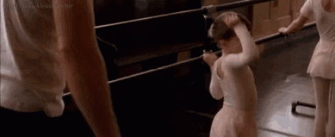a little girl in a pink tutu is standing next to a bar in a dance studio .