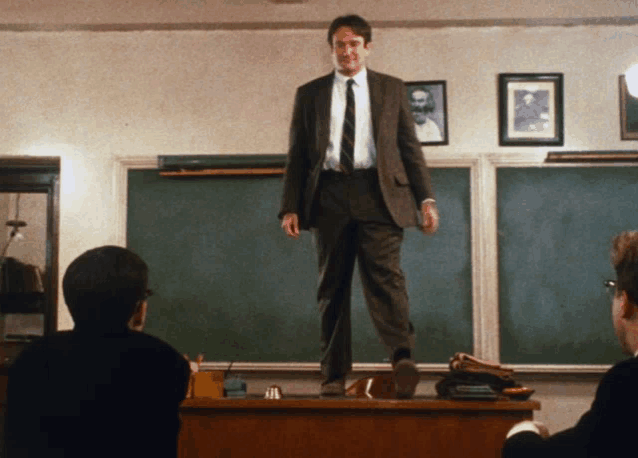 a man in a suit and tie stands on a desk in a classroom
