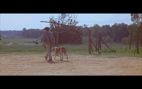 a man walking a dog on a dirt road with a fence in the background